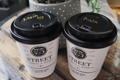 Two takeaway coffee cups labeled with women’s names on a rustic wooden table beside a small potted plant, symbolizing friendship, connection, and shared moments between women.
