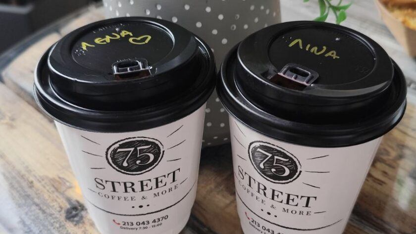 Two takeaway coffee cups labeled with women’s names on a rustic wooden table beside a small potted plant, symbolizing friendship, connection, and shared moments between women.