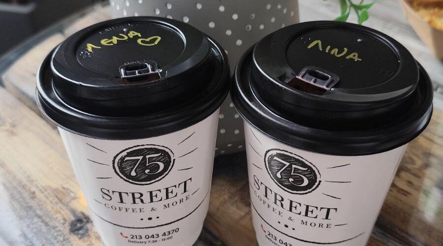 Two takeaway coffee cups labeled with women’s names on a rustic wooden table beside a small potted plant, symbolizing friendship, connection, and shared moments between women.